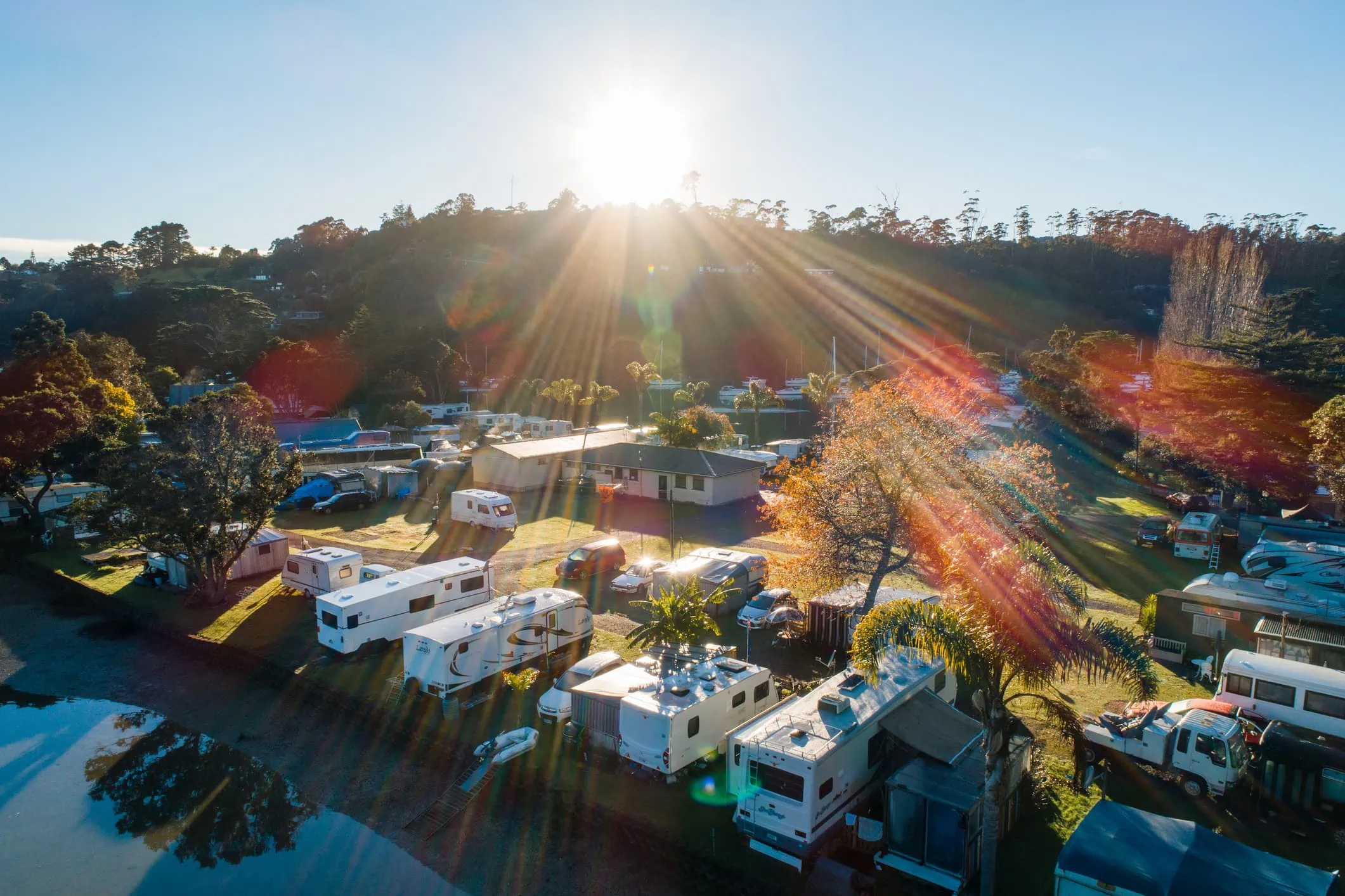 A scenic view of a campsite at sunrise with several RVs and trailers parked under a clear sky. Sun rays illuminate the area, casting light over trees and a body of water nearby.