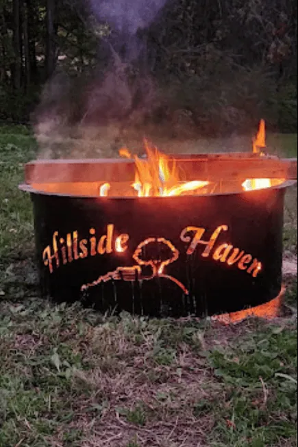 A metal fire pit with flames and smoke rising. The fire pit is engraved with "Hillside Haven" and an image of a tree. It's situated outdoors on grass with trees visible in the background.
