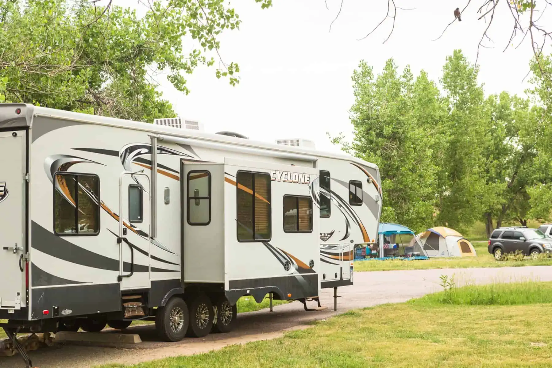 A large white RV with abstract black and gray designs is parked on a grassy area near a campsite. Tents and a parked car are visible in the background among green trees under a cloudy sky.