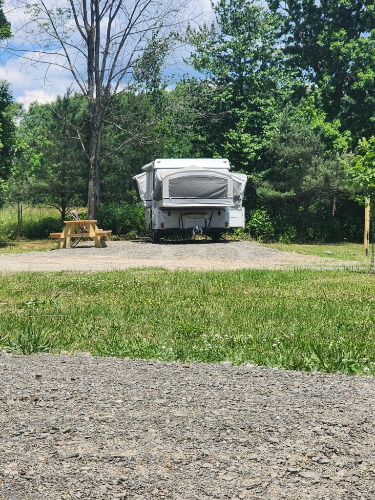 A pop-up camper is set up in a grassy area surrounded by trees. A wooden picnic table sits to the left of the camper. The sky is partly cloudy, creating a serene outdoor atmosphere.
