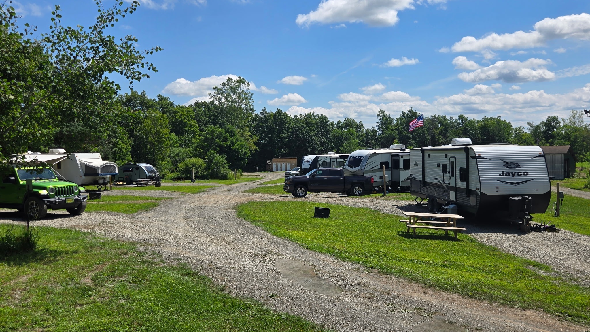 Several RVs and campers are parked on grassy lots along a gravel road at a campground. A green Jeep and a pickup truck are visible; a picnic table sits on the grass. Trees and blue sky with clouds are in the background.